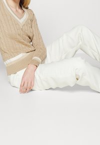 Beige kabelstickad tröja med V-ringning och vita accenter, i kombination med krämfärgad byxor som är uppdragna vid anklarna. Mjuk textur, avslappnad passform.