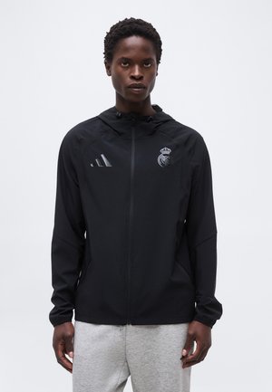 Νέος άνδρας που φοράει μαύρο μπουφάν με φερμουάρ και κουκούλα της Ρεάλ Μαδρίτης και γκρι φόρμα, στέκεται μπροστά σε απλό λευκό φόντο.