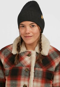 Sort ribbet beanie og en rød ternet jakke med en krave af cremet kunstpels, der har runde brune knapper og et rutet mønster.