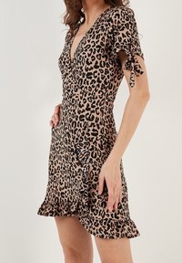 Donna che indossa un vestito avvolgente corto con stampa leopardata, con orlo a volants e dettaglio con fiocco sulle maniche corte, in piedi di fronte a uno sfondo neutro.