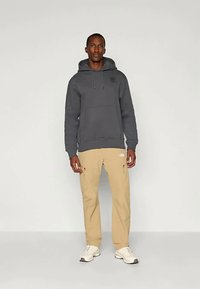 Άνδρας στέκεται μπροστά σε απλή φωτεινή επιφάνεια, φορώντας γκρι φούτερ, μπεζ cargo παντελόνι με φερμουάρ, και λευκά αθλητικά παπούτσια.