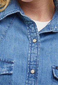 Helles, blaues Jeanshemd mit Druckknöpfen und strukturiertem Kragen, zwei vorderen Taschen und subtilen Texturdetails.