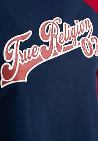Blaues T-Shirt mit roten und weißen Akzenten, das ein großes „True Religion 02“-Logo in stilisierter Schriftart zeigt und aus weichem Baumwollmaterial besteht.