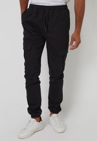 Schwarze Cargo-Hosen mit elastischem Bund, zwei seitlichen Taschen und gerippten Bündchen; glatte Textur und leichtes Material. Getragen mit weißen Sneakern.
