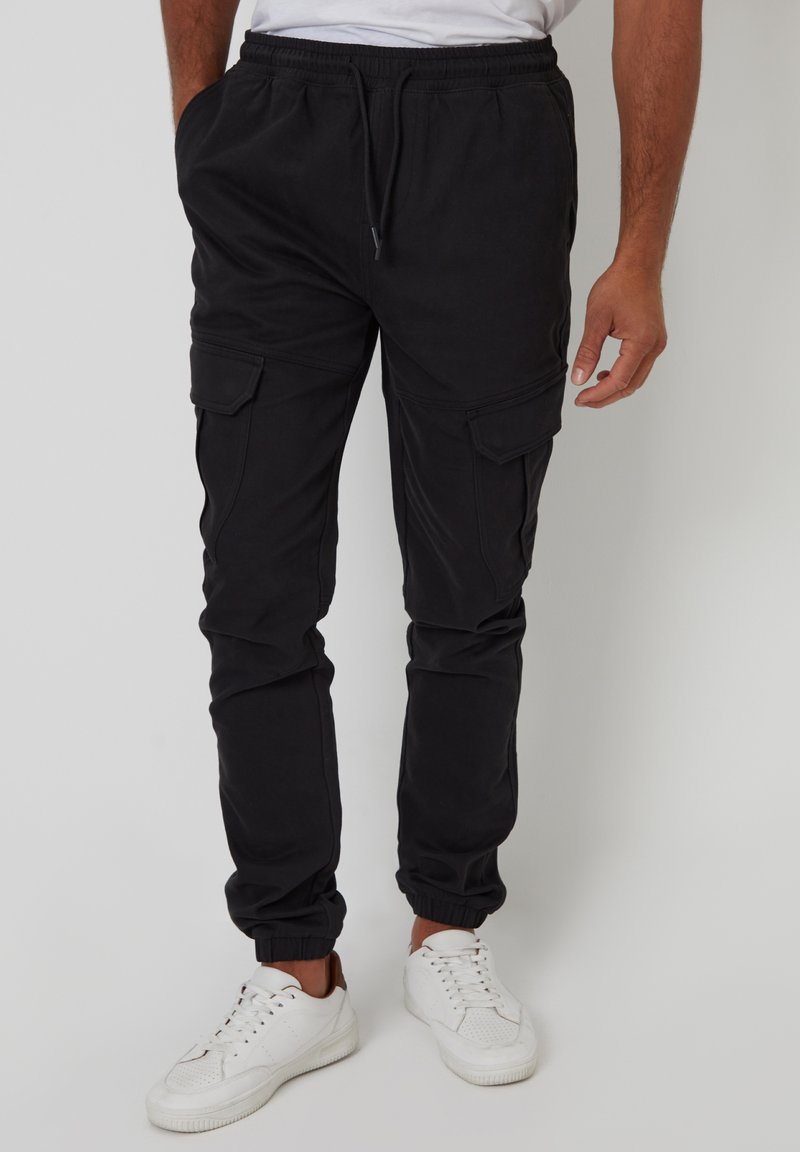 Schwarze Cargo-Hosen mit elastischem Bund, zwei seitlichen Taschen und gerippten Bündchen; glatte Textur und leichtes Material. Getragen mit weißen Sneakern.