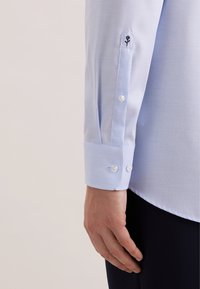 Lichtblauw overhemd met een textuurstof. Heeft een puntige manchetknoop met twee knopen en een geborduurd bloemdetail vlak bij de manchet.