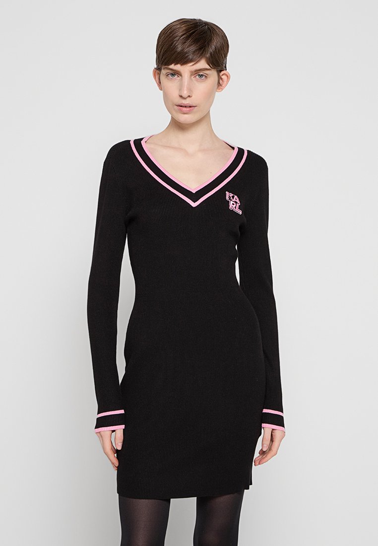 Frau in einem schwarzen langärmeligen Strickkleid mit rosa Besatz am V-Ausschnitt, den Bündchen und dem "KARL"-Logo auf der Brust, steht vor einem schlichten Hintergrund.