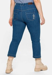 Mörkblå jeans i denim med avsmalnande ben, med uppvikta fållar och blommönsterbroderi på bakfickorna. Gyllene öglor och läderpatchdetalj.