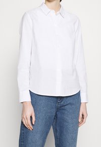 Vit långärmad skjorta med knappar och krage, med en slät textur och rak fåll, bärs över blå denimjeans.