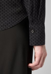 Zbliżenie na rękę spoczywającą na boku, ubraną w czarną koszulę w wzór houndstooth z zapinanym mankietem.