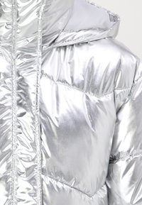 Chaqueta puffer plateada con un acabado brillante, diseño acolchado, gran capucha y detalles de costura visibles. Textura suave, apariencia ligera.