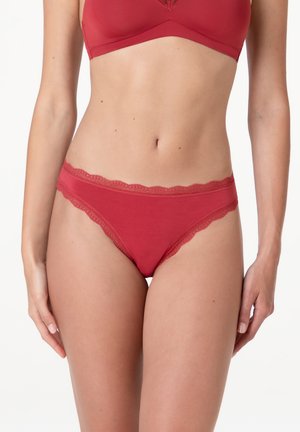 Donna che indossa mutandine rosse con bordatura in pizzo e reggiseno coordinato, mostrata dalla metà coscia fino sotto il petto su sfondo bianco.
