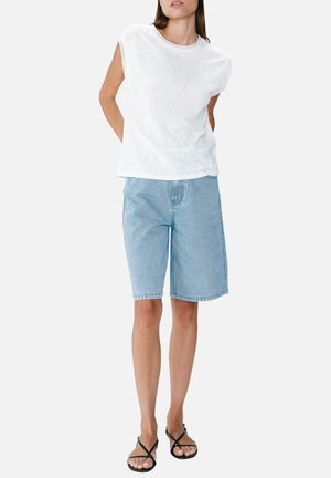 Femme portant un haut blanc sans manches, un short en jean bleu clair longueur genoux et des sandales noires à brides, debout devant un fond uni.