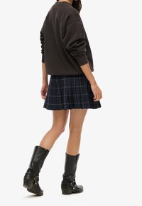 Sudadera negra con hombros caídos, falda plisada de cuadros azul marino y botas negras de cuero hasta la rodilla con hebillas.