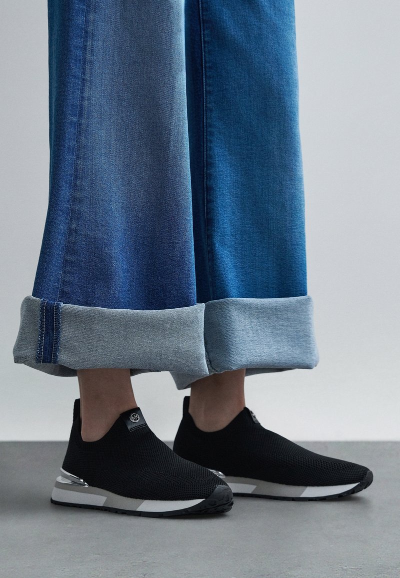 Personne portant un jean bleu à jambes larges retroussé aux chevilles et des baskets slip-on noires avec semelles blanches, debout sur un sol gris.