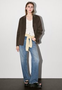 Brown blazer, white t-shirt, yellow sweater tied at the waist, light blue flared jeans, and black platform sandals. Plain background.
