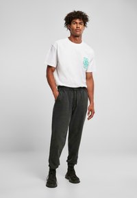 Hvid t-shirt med et turkist logo på venstre bryst, parret med sorte joggerbukser med elastisk talje og opslåede kanter.