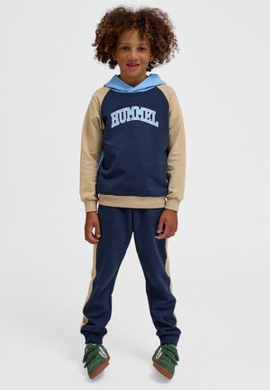 Barn med krøllet hår iført en blå og beige Hummel hættetrøje og matchende bukser, stående og smilende, iført grønne sneakers på hvid baggrund.