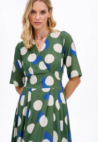 Zelené šaty s překládaným designem, zdobené velkými bílými a modrými puntíky. Krátké rukávy a mírně rozšířená sukně. Hladká textura látky.