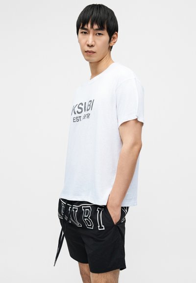 Άσπρη βαμβακερή μπλούζα με μαύρη εκτύπωση "KSUBI EST." μπροστά, συνδυασμένη με μαύρο σορτς με λευκές λογότυπες λεπτομέρειες και κορδόνι στη μέση.