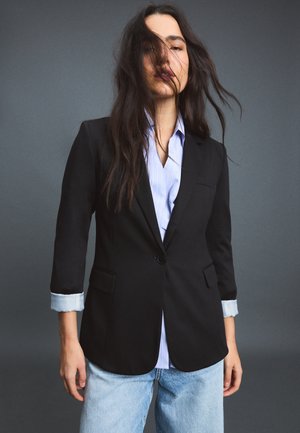 Femme aux longs cheveux foncés porte un blazer noir sur une chemise bleu clair et un jean, debout devant un fond gris. Les cheveux couvrent partiellement son visage.