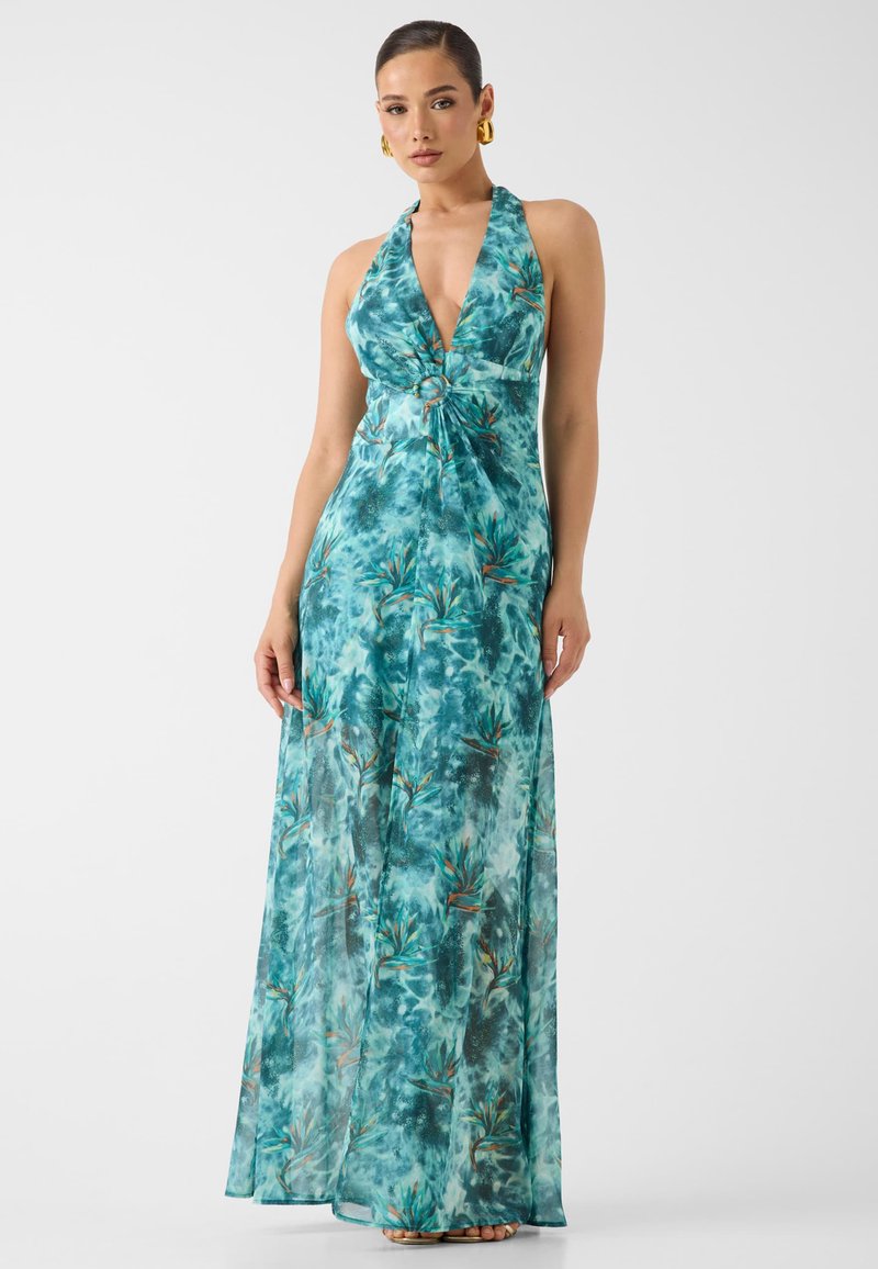 Femme portant une robe longue dos nu bleu sarcelle et verte avec un motif floral et un détail anneau à l'avant, debout devant un fond uni.