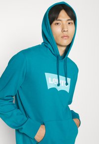 Turkooise hoodie met een kangaroo zak, verstelbare koord en een wit Levi's logo op de voorkant; gemaakt van zachte stof.