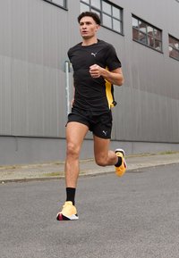 Maglietta sportiva nera con accenti gialli, pantaloni corti neri e scarpe da corsa arancioni chiare con dettagli rosa e neri. Corse su pavimentazione.