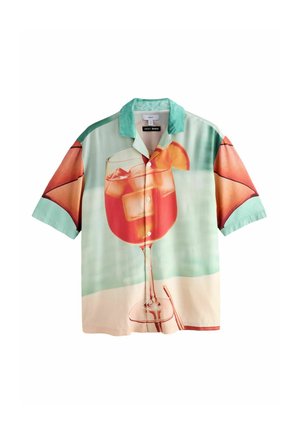 Young man standing outdoors against beige wall wearing colorful short-sleeve shirt with large drink print, pink striped shorts, and white sneakers.