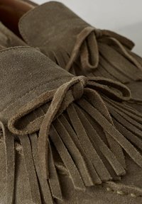 Mocasines de ante marrón con un diseño cosido y detalles de flecos en la parte delantera, complementados con borlas a juego y un acabado suave.
