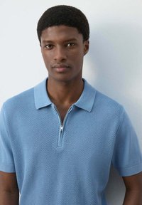 Joven con el pelo corto y rizado, que lleva una camisa polo de manga corta azul claro con textura y una cremallera parcial, posando frente a un fondo blanco liso.