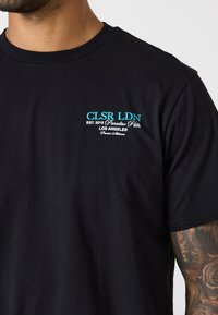 Svart bomull T-skjorte med turkise tekster og merkevaredetaljer. Korte ermer og standard rund halsåpning. Jevn tekstur.