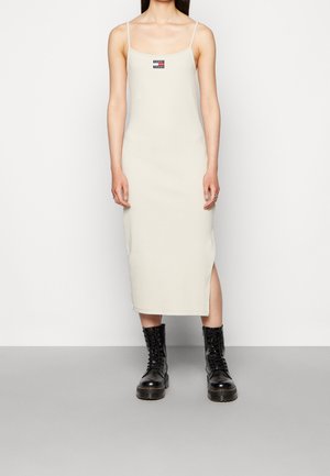 Crème midi-jurk met dunne bandjes, voorzien van zijsplitten en een klein rood, wit en blauw logo-opdruk. Gecombineerd met zwarte veterschoenen.