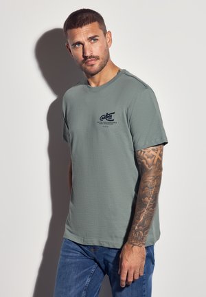 Man with short hair and arm tattoo wearing a gray T-shirt and blue jeans, standing against a white wall casting a shadow.