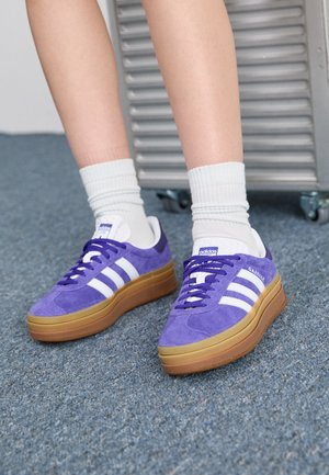 Nogi w białych skarpetkach i fioletowych sneakersach Adidas Gazelle z gumowymi podeszwami, stojące na niebieskim dywanie obok metalowego wylotu powietrza na kółkach.