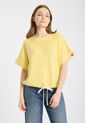 Młoda kobieta o brązowych włosach do ramion, ubrana w luźną żółtą bluzkę z krótkim rękawem, białym sznurkiem i niebieskie dżinsy na białym tle.