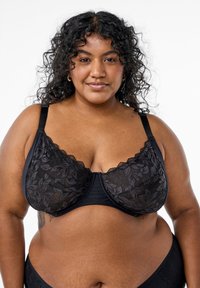 Reggiseno di pizzo nero con motivi floreali, spalline regolabili e bordi ondulati. Il tessuto è liscio con un pannello superiore semi-trasparente.