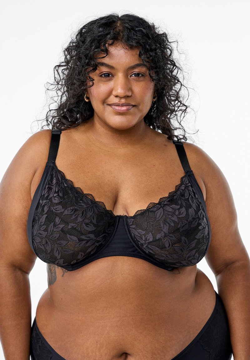 Reggiseno di pizzo nero con motivi floreali, spalline regolabili e bordi ondulati. Il tessuto è liscio con un pannello superiore semi-trasparente.