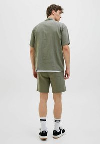 Grøn, struktureret kortærmet skjorte parret med matchende shorts. Båret med sorte sneakers og hvide stribede sokker. Ensfarvet baggrund.