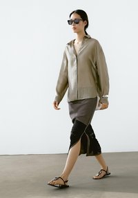 Lichtgrijs overhemd met knopen, zwarte rok met geplooide textuur en platte zwarte sandalen. De omgeving heeft neutrale tinten en minimale decoratie.