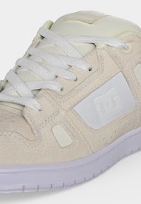 Κοντινό πλάνο μπεζ και λευκού sneaker DC με υφασμάτινες σουέτ και διχτυωτές επιφάνειες, λευκά κορδόνια και ανάγλυφο λογότυπο DC στο πλάι.