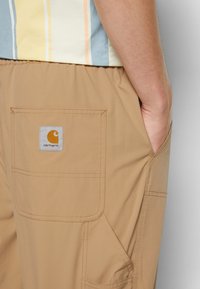 Béžové cargo šortky s boční kapsou a vyšitým logem Carhartt. Tkanina se zdá být lehká s hladkou texturou.