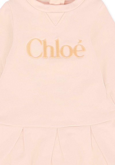 Robe rose clair en tissu doux avec un décolleté en V, une jupe plissée, et "Chloé" brodé en beige clair sur le devant.