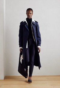 Navy trench coat with white collar detail, paired with a black turtleneck and slim trousers. Carrying a black bag, wearing black open-toe shoes.