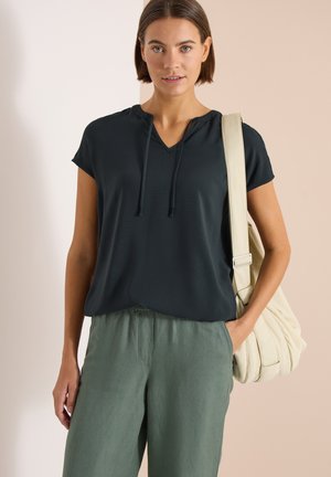 Donna che indossa un top nero a maniche corte con scollo a V, pantaloni verdi, e porta una borsa a tracolla imbottita beige con una mano in tasca.