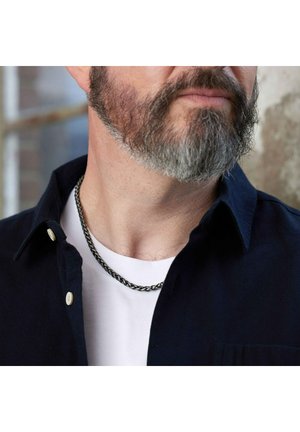 Zilverkleurige metalen ketting, plat en geweven ontwerp, gedragen over een wit T-shirt onder een donker overhemd met een lichte kraag en beige knopen.