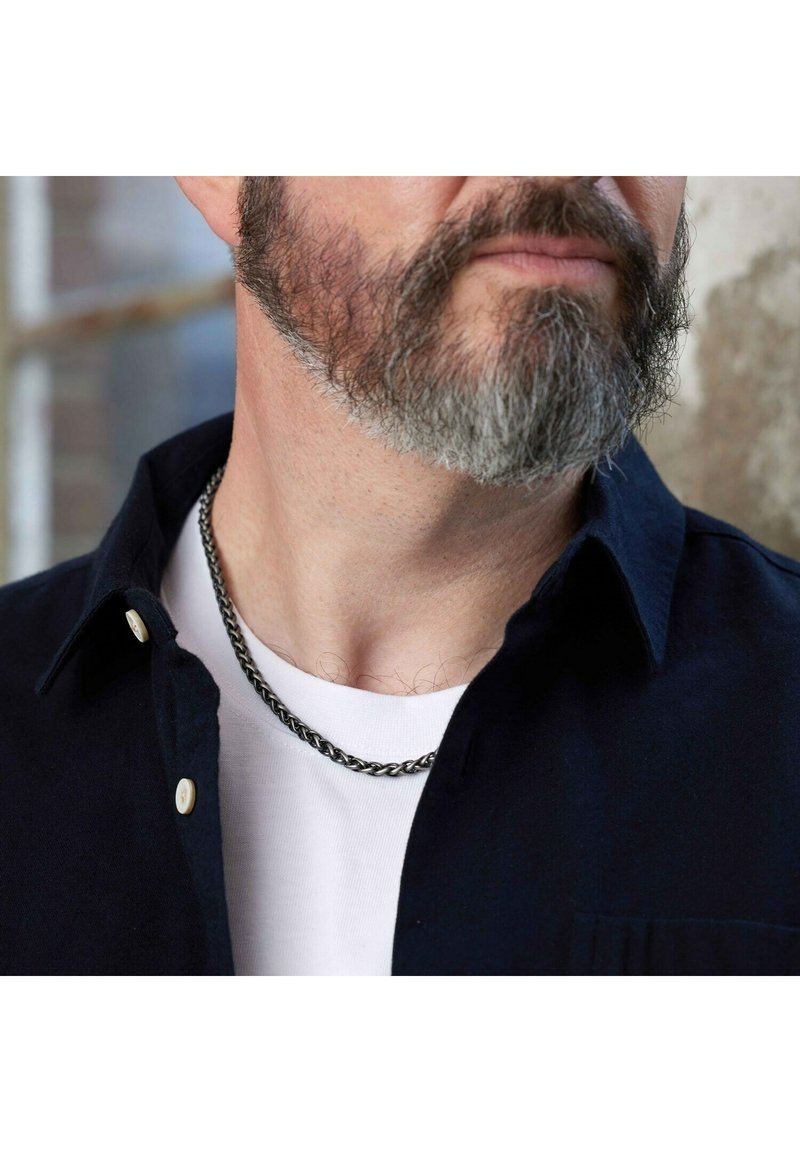 Zilverkleurige metalen ketting, plat en geweven ontwerp, gedragen over een wit T-shirt onder een donker overhemd met een lichte kraag en beige knopen.