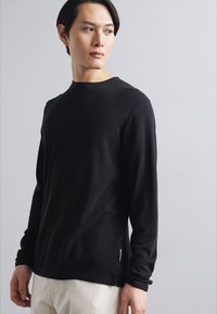Schwarzes Langarmshirt aus glattem Stoff mit Rundhalsausschnitt, seitlichen Schlitzen und minimalen Nahtdetails, kombiniert mit hellen Hosen.