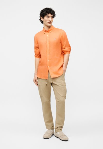 Άνδρας με σγουρά σκούρα μαλλιά που φοράει πορτοκαλί πουκάμισο με κουμπιά και μανίκια γυρισμένα, μπεζ παντελόνι cargo και ανοιχτό μπεζ παπούτσια, στέκεται μπροστά σε απλό φόντο.