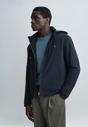 Mladi moški v temno modri jakni z zadrgo, modri potiskani majici in bež hlačah, stoji z eno roko v žepu in gleda levo na enobarvnem ozadju.